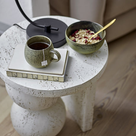 Tally Round Resin Side Table in white with pitted stone-like texture, shown bedside holding a tea bag, mug, notebook, and ceramic bowl of oats.