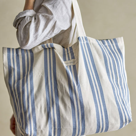 Trina Striped Cotton Shopping Bag in blue, carried over the shoulder, showing vertical stripes, long fabric handles and a fabric brand label on the front.