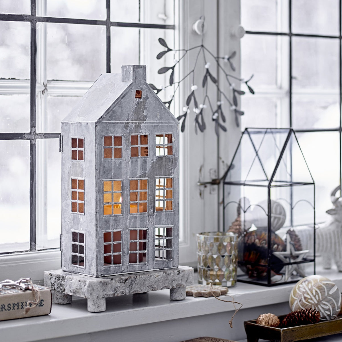 Grey iron house-shaped Christmas lantern with pitched roof, chimney and paned windows lit from within, displayed on a wooden stand by a window.