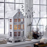Grey iron house-shaped Christmas lantern with pitched roof, chimney and paned windows lit from within, displayed on a wooden stand by a window.