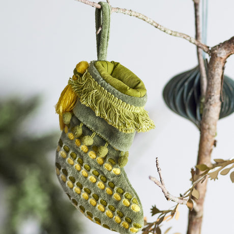 Jeanne Cotton Christmas Tree Decoration in green, a small padded stocking with a fringed cuff, yellow tassel and pom-pom trim, and dotted foot pattern, hanging from a branch.