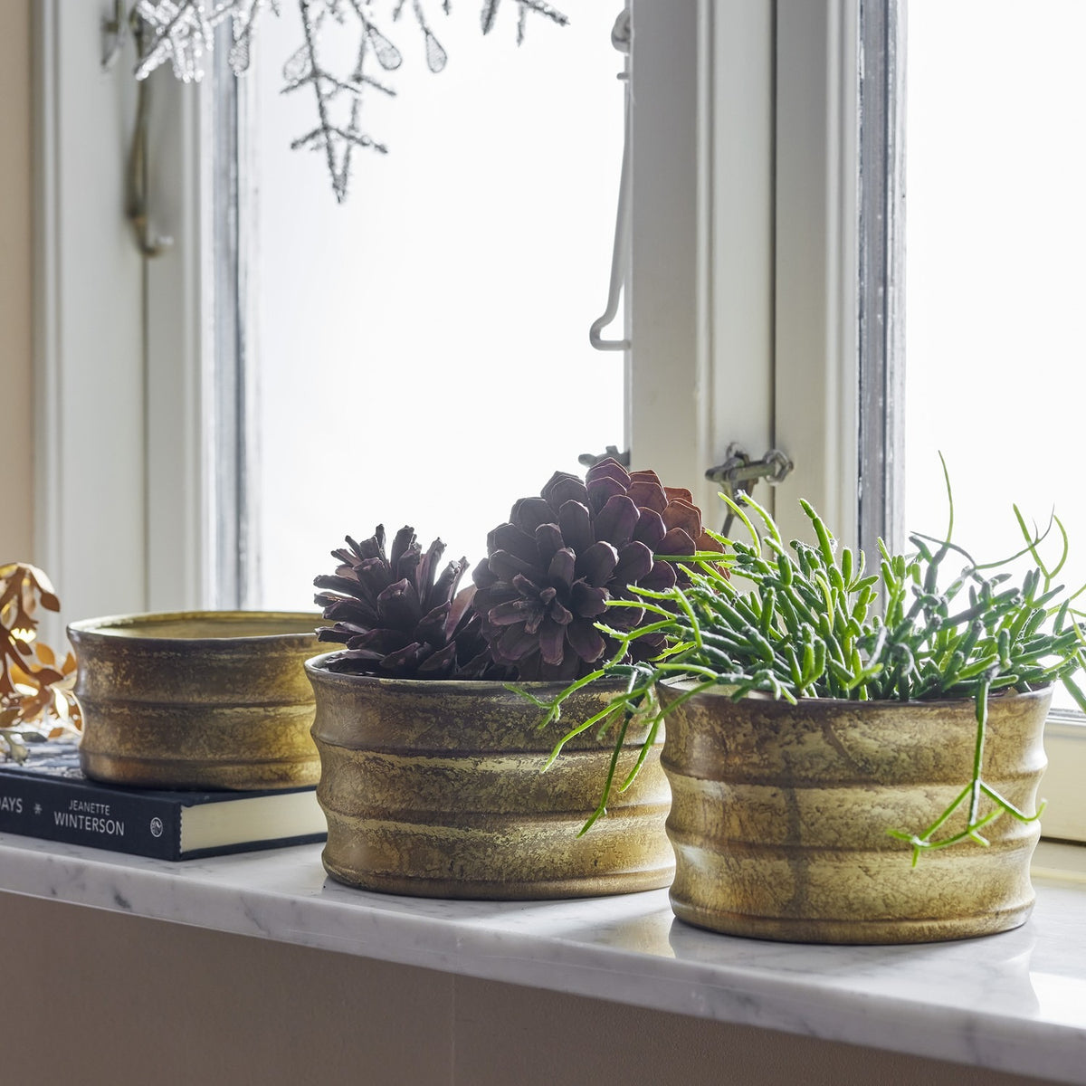 Lucilla Iron Christmas Plant Pots in worn gold, ribbed round design, shown on a marble windowsill holding pinecones and trailing greenery.