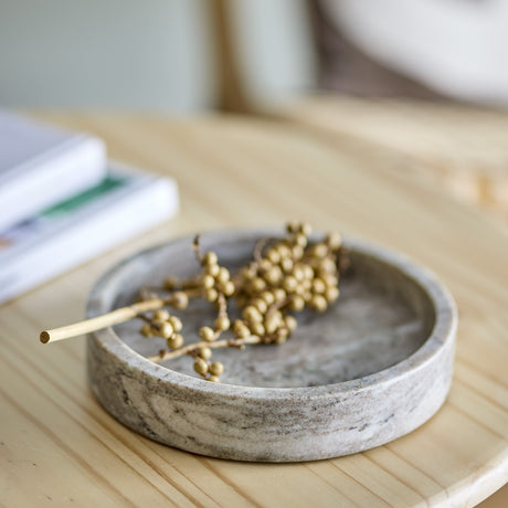 Ivette Round Marble Tray in grey with a low, shallow design and upright rim, shown on a wooden table holding a gold berry sprig.