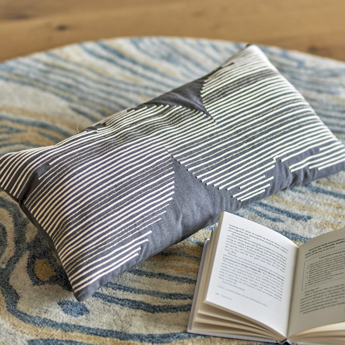 Brechia Striped Cotton Cushion in grey, rectangular with embroidered stripe pattern forming abstract shapes, shown on a patterned rug beside an open book.