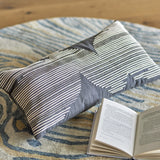 Brechia Striped Cotton Cushion in grey, rectangular with embroidered stripe pattern forming abstract shapes, shown on a patterned rug beside an open book.