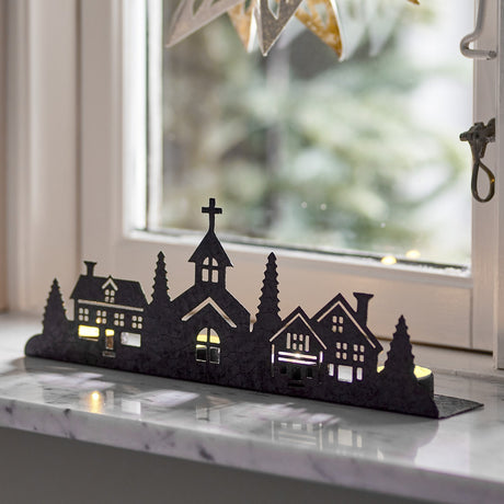 Cielo Iron Christmas Tealight Holder in black, a village silhouette of houses, fir trees and a church with cutout windows, shown lit on a marble windowsill.