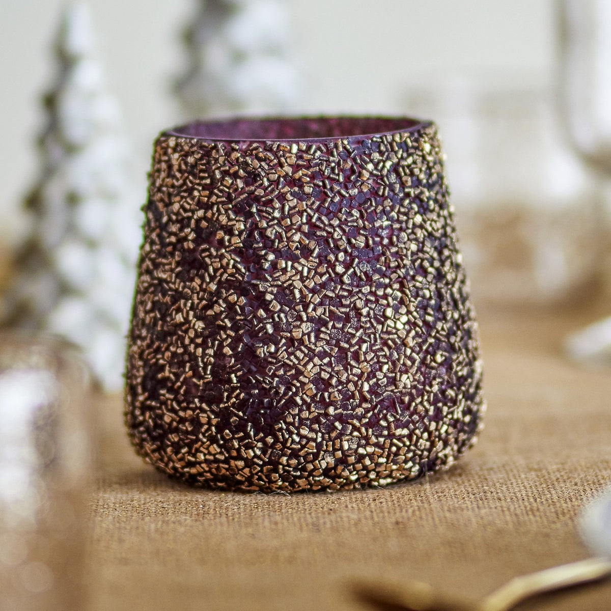 Filiz Glass Christmas Tealight Holder in Gold, round recycled glass votive with dense textured gold mosaic finish, tapered shape, shown on a burlap surface with festive decor in the background.