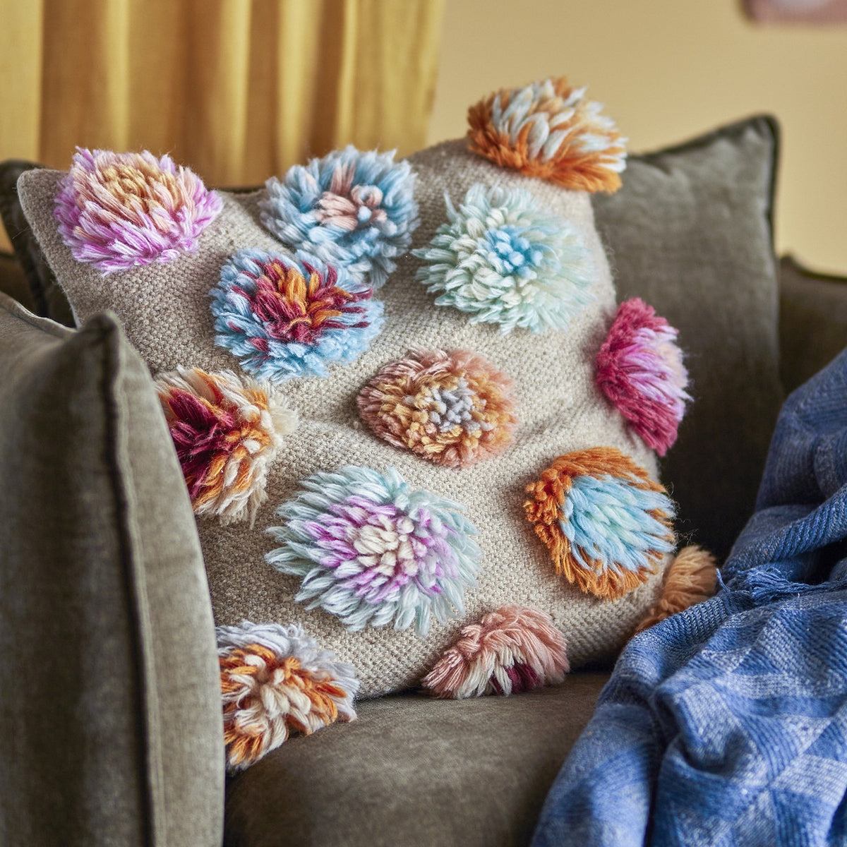 Somma Wool Cushion in Multi with shaggy tufted flower-like yarn clusters on a woven beige base, shown on a sofa with a blue throw.
