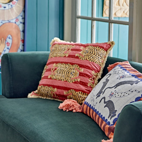 Poirino Cotton Cushion in pink with a striped tiger print and fringed edges, shown on a dark green sofa beside a patterned cushion.