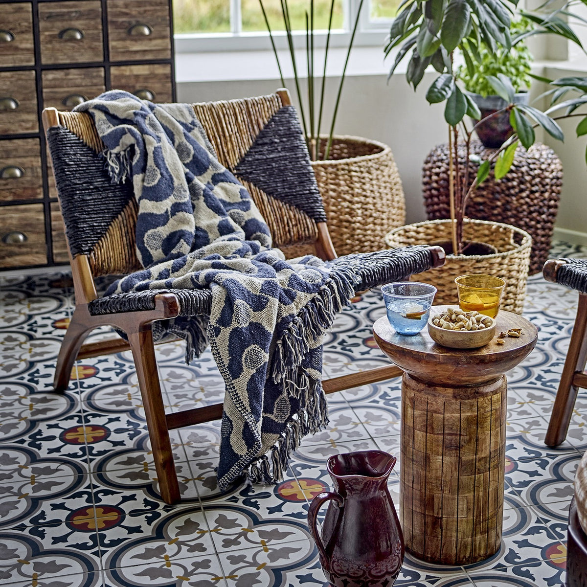 Santena Wavy Cotton Throw in blue with fringed edges, draped over a woven wooden armchair in a living room with patterned tiled flooring.