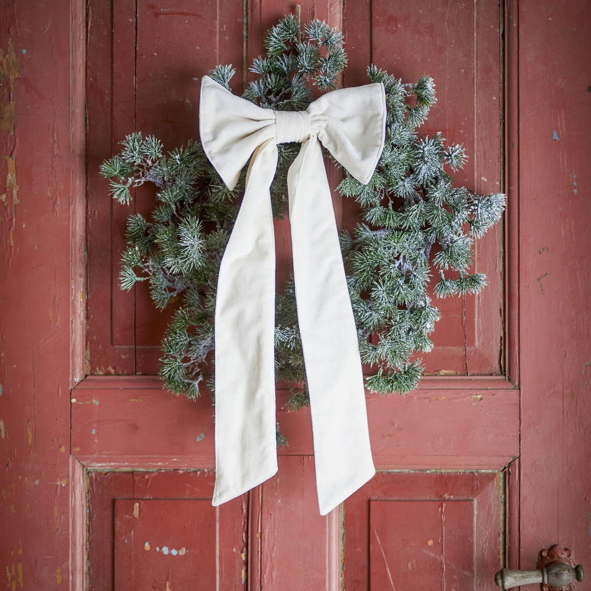 Ruby cream cotton Christmas bow with long trailing tails, fastened to a frosted pine wreath hanging on a weathered red door.