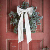 Ruby cream cotton Christmas bow with long trailing tails, fastened to a frosted pine wreath hanging on a weathered red door.