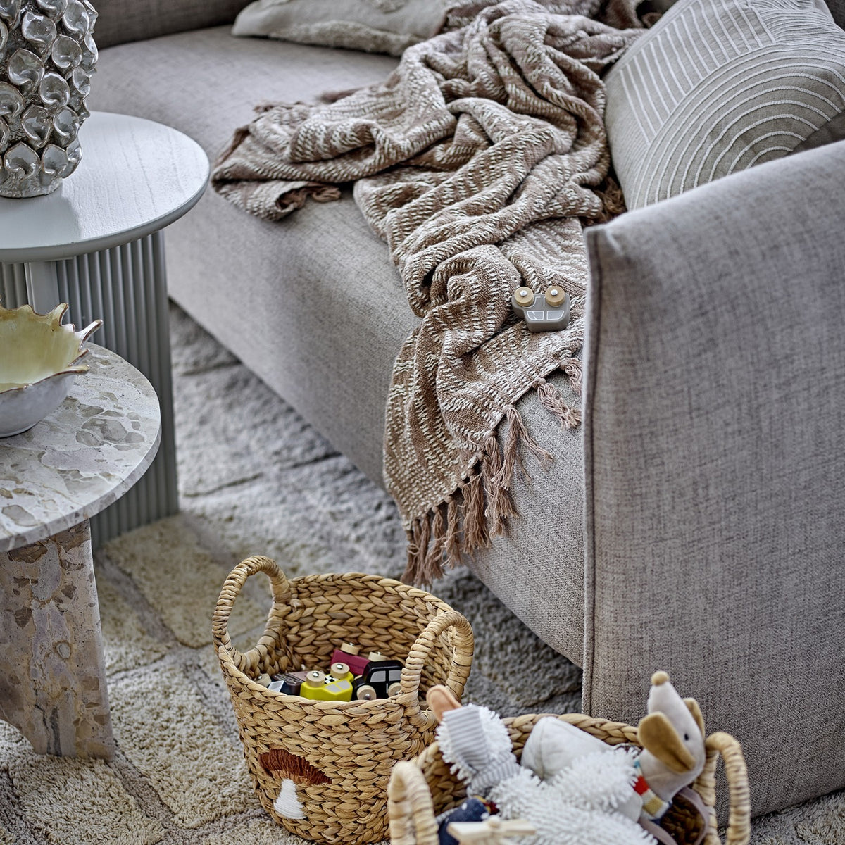 Okapi Cotton Throw in beige with woven wavy-line texture and fringed edge, draped over a grey sofa beside woven toy baskets.