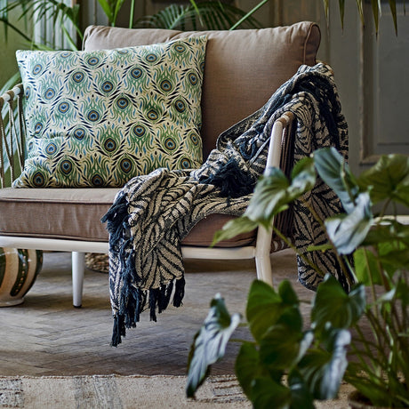 Ugento Leaf Cotton Throw in green, woven leaf pattern with knotted fringe, draped over a brown armchair beside a patterned cushion in a plant-filled living room.