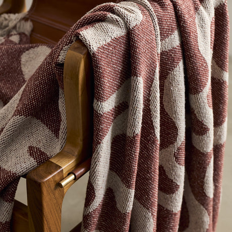 Salobre Cotton Throw in brown with a woven abstract organic pattern, draped over the wooden arm of a chair, showing its textured cotton-blend weave.