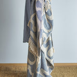 Salobre Cotton Throw in blue with abstract cream pattern and knotted fringe edges, draped over a chair against a plain light grey background.