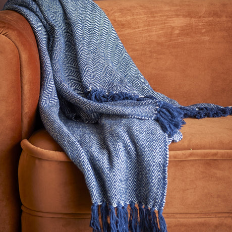 Herringbone Throw in blue with fringed edges, draped over a tan velvet sofa, showing its zigzag woven pattern and tasselled trim.