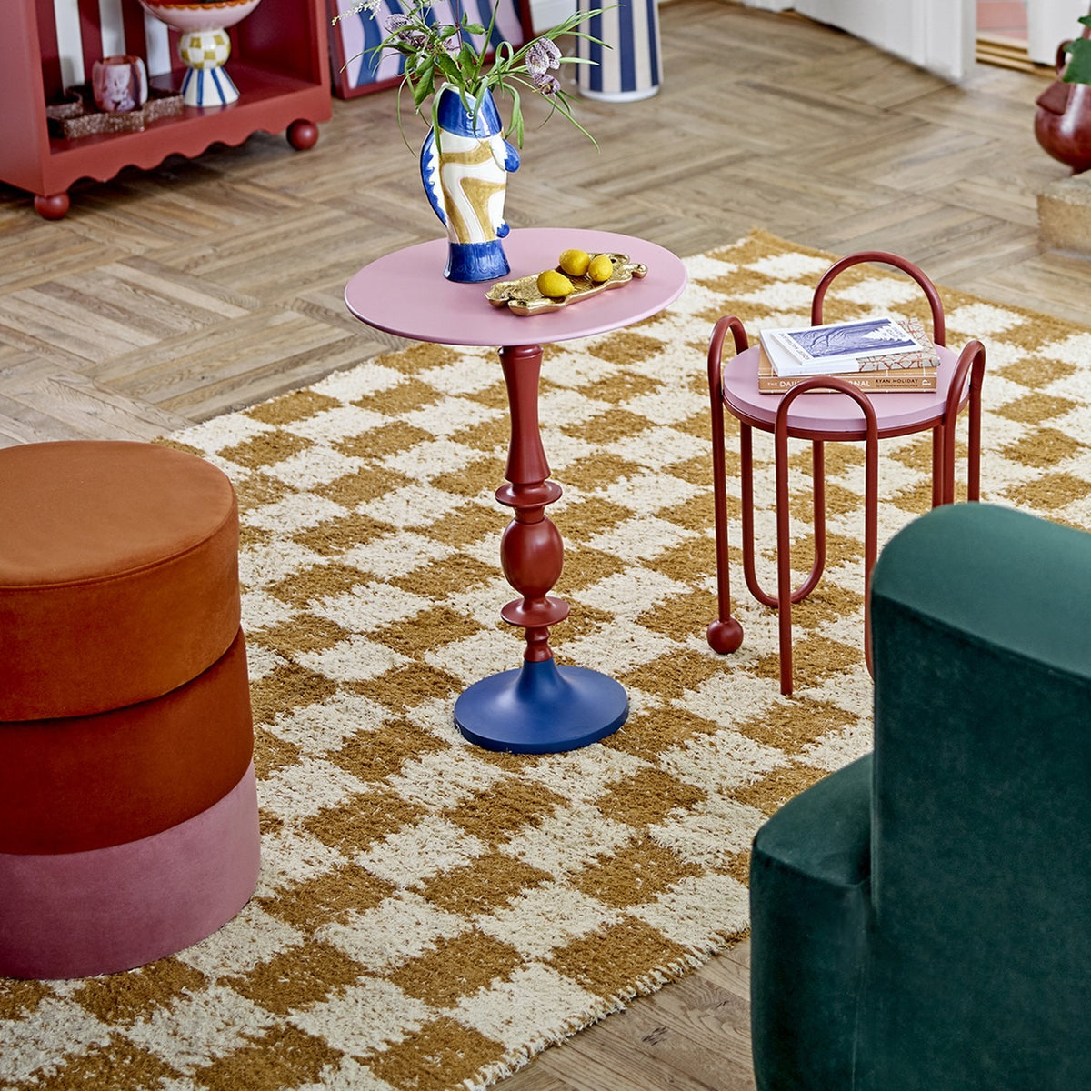 Chola Cotton Rug 200x140cm in brown, shown in a living room with a woven checkerboard pattern beneath a pedestal side table, trolley, pouffes and green armchair.