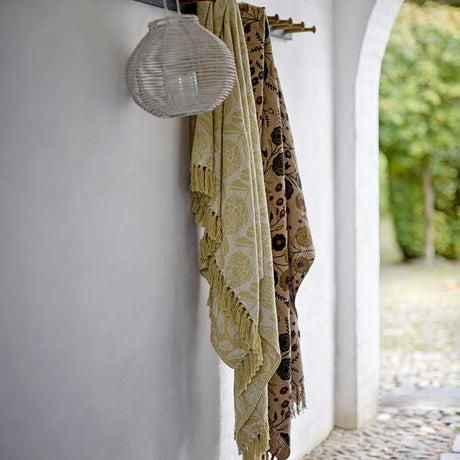 Pratello Throw in yellow with a woven floral pattern and fringed edge, draped over a hook beside a patterned brown throw in an outdoor archway setting.