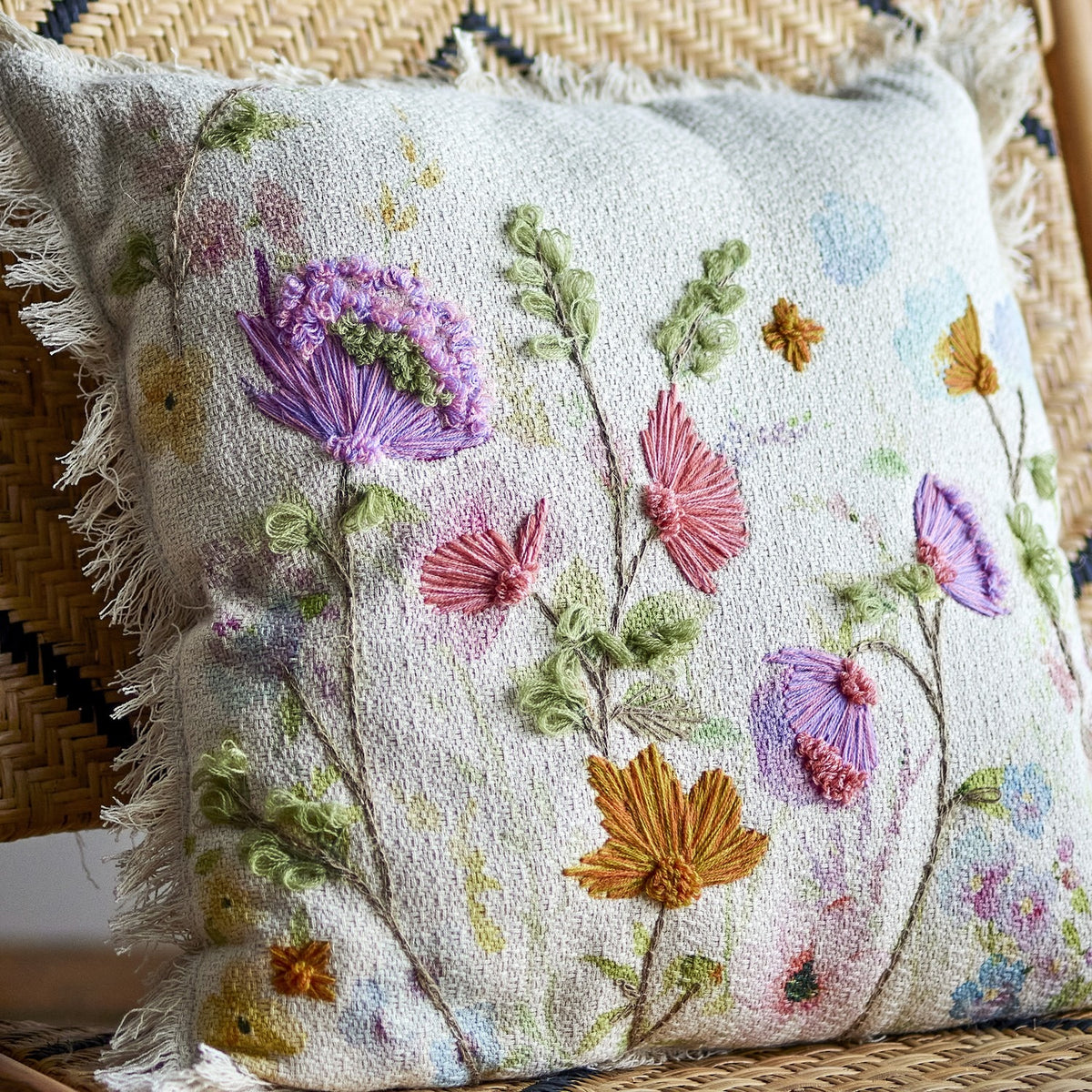 Pogni Floral Cotton Cushion in Natural, square with fringed edges, showing raised embroidered wildflowers in purple, pink and orange over a watercolour-style floral print, shown on a woven chair.