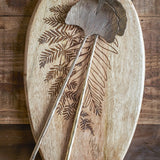 Lyas Iron Salad Servers Set of 2 in brass, ginkgo leaf-shaped heads with ribbed fan detail, resting on an engraved oval wooden board.