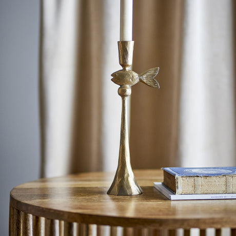 Capet Iron Candle Holder in brass finish, featuring a fish figure below the candle cup and a slender stem on a bell-shaped foot, shown on a wooden table with a book nearby.