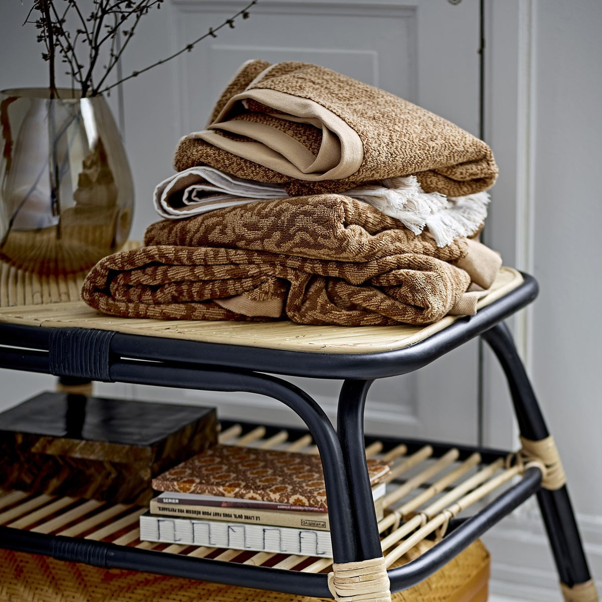 Stack of folded Kaysa cotton hand towels in brown patterned and solid cream tones, displayed on a rattan shelf unit with books and a vase below.