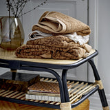 Stack of folded Kaysa cotton hand towels in brown patterned and solid cream tones, displayed on a rattan shelf unit with books and a vase below.