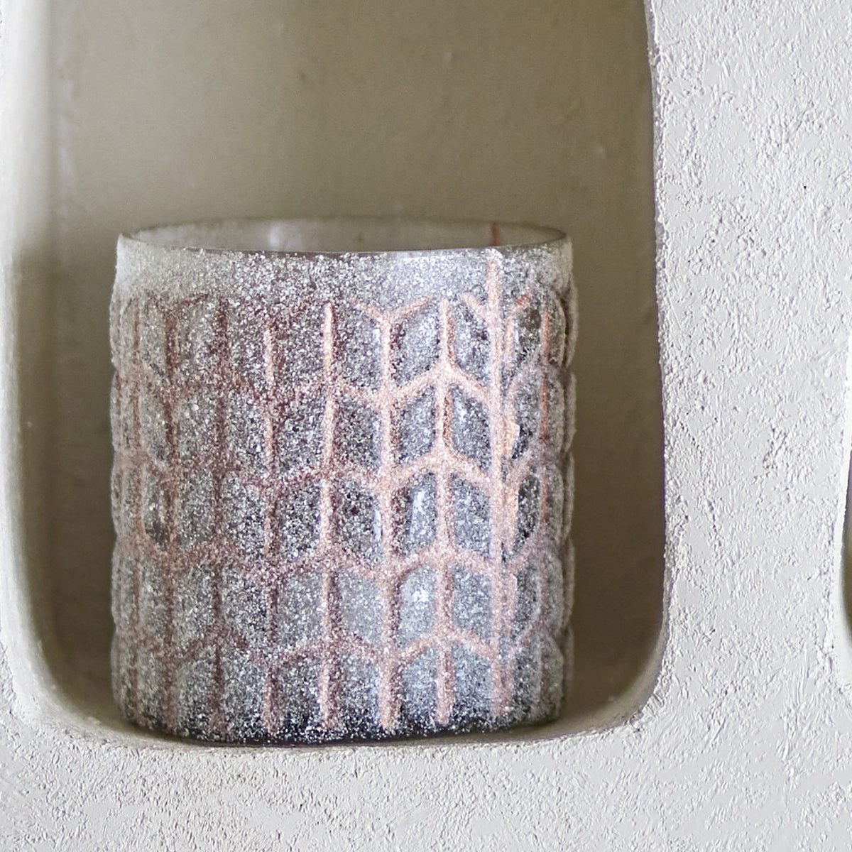 Esther Glass Tealight Holder in pink, a cylindrical holder with a frosted granular texture and raised chevron lattice pattern, displayed in a wall niche.