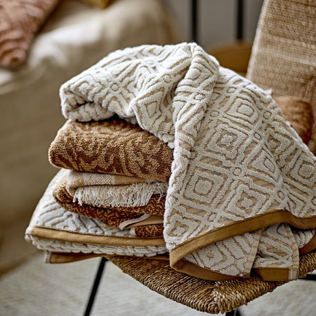 Malou Cotton Towel in Natural, folded with a raised diamond and square pattern, stacked among brown-toned towels on a woven chair.