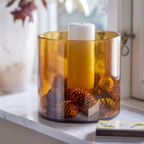 Kahlah amber glass candle holder with white pillar candle and pinecones, displayed on a marble windowsill.
