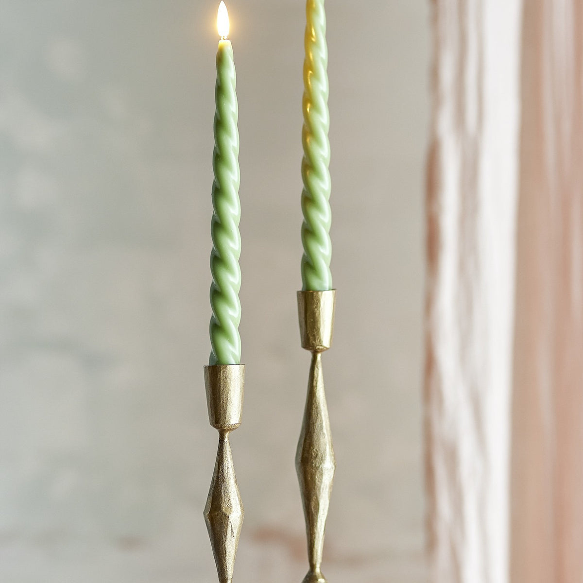 Ponza LED taper candles, pack of 2, green spiral-twist wax body with flame-shaped tip, shown lit in brass candle holders against a softly blurred interior backdrop.