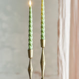 Ponza LED taper candles, pack of 2, green spiral-twist wax body with flame-shaped tip, shown lit in brass candle holders against a softly blurred interior backdrop.