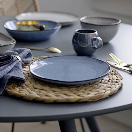 Sandrine blue stoneware dinner plate with reactive glaze on a woven placemat, set alongside a matching mug, bowl, deep plate and gold cutlery on a grey dining table.