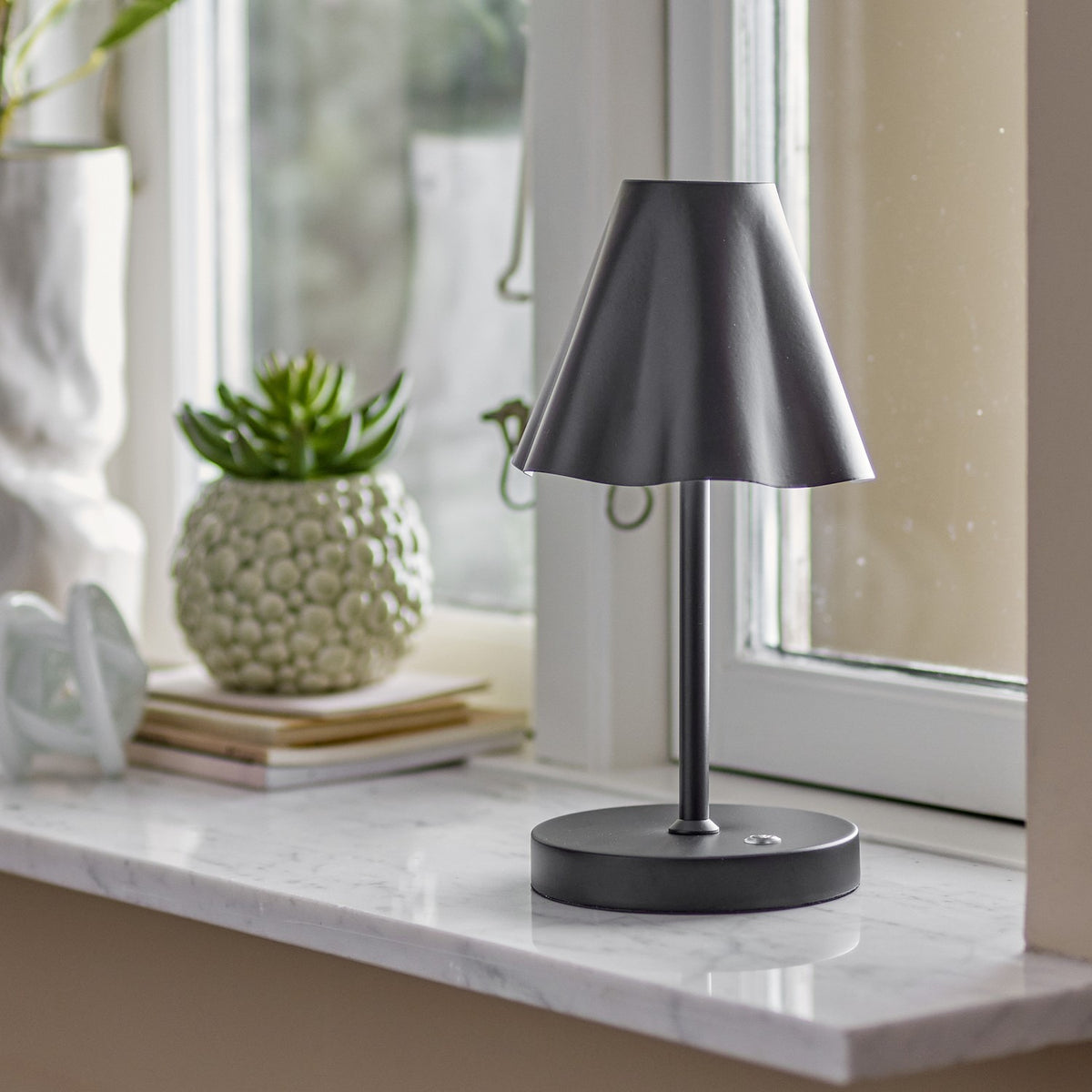 Lianna Wavy Iron Rechargeable Table Lamp in black, with a cone-shaped wavy-edge shade on a slender stem and round base, shown on a marble windowsill.