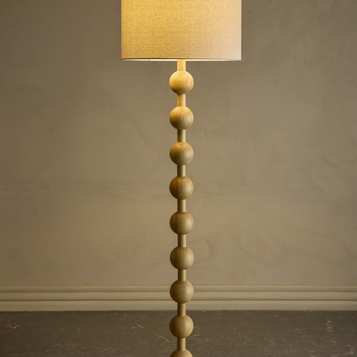 Giacoma Wooden Floor Lamp in natural wood, featuring a stacked sphere stem and a tall linen drum shade, lit against a plain grey background.