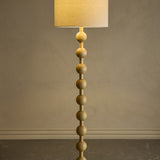 Giacoma Wooden Floor Lamp in natural wood, featuring a stacked sphere stem and a tall linen drum shade, lit against a plain grey background.