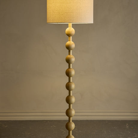 Giacoma Wooden Floor Lamp in natural wood, featuring a stacked sphere stem and a tall linen drum shade, lit against a plain grey background.