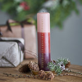 Dip Dye Christmas Candle advent pillar in paraffin wax, dip-dyed in pink, red and burgundy bands with gold numbers 1 to 24, shown on a wooden table with pinecones and gift boxes.