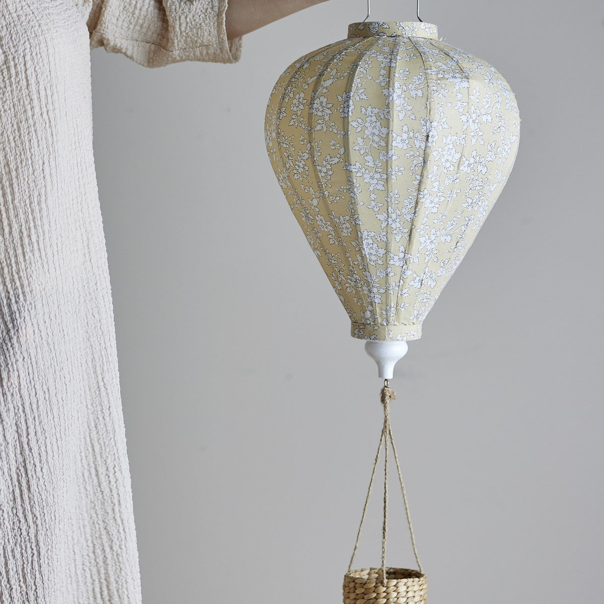 Yellow hot air balloon-shaped baby mobile with small floral pattern, white finial, and woven basket on cords, held in hand against a plain light background.