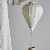 Yellow hot air balloon-shaped baby mobile with small floral pattern, white finial, and woven basket on cords, held in hand against a plain light background.