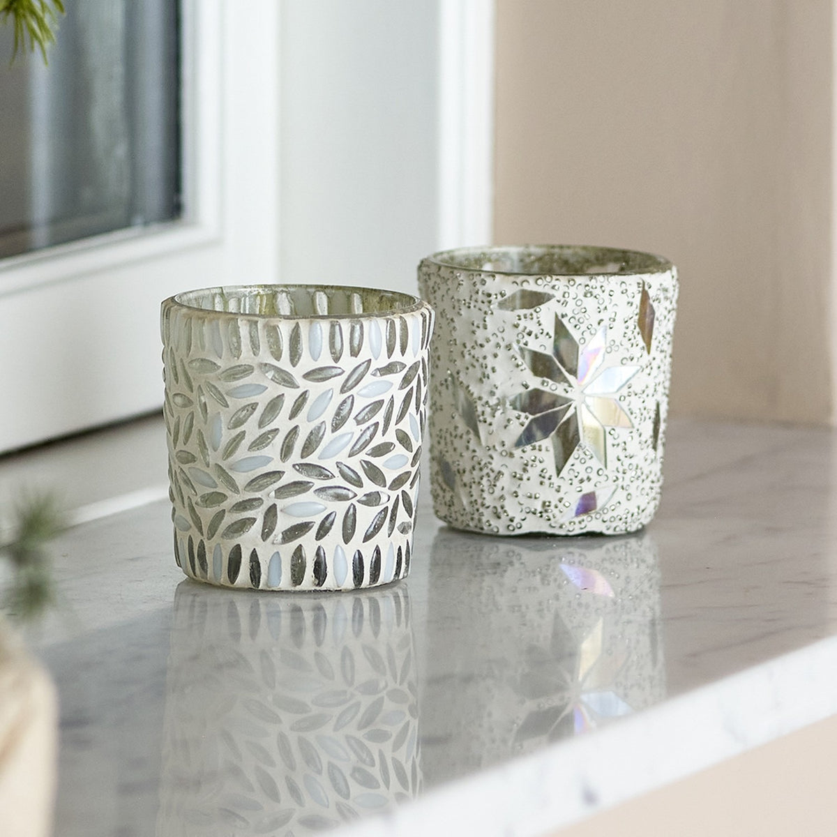 Myr glass tealight holders, set of 2, white mosaic design with leaf pattern and star motif, shown on a marble surface by a window.