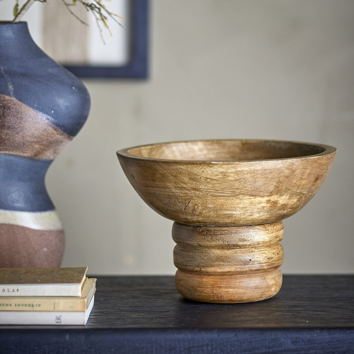 Nanuka Round Wooden Pedestal Bowl 20.5cm in Brown, mango wood with visible grain, on a ribbed stacked-ring foot, shown on a dark shelf beside books and a patterned vase.