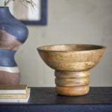 Nanuka Round Wooden Pedestal Bowl 20.5cm in Brown, mango wood with visible grain, on a ribbed stacked-ring foot, shown on a dark shelf beside books and a patterned vase.