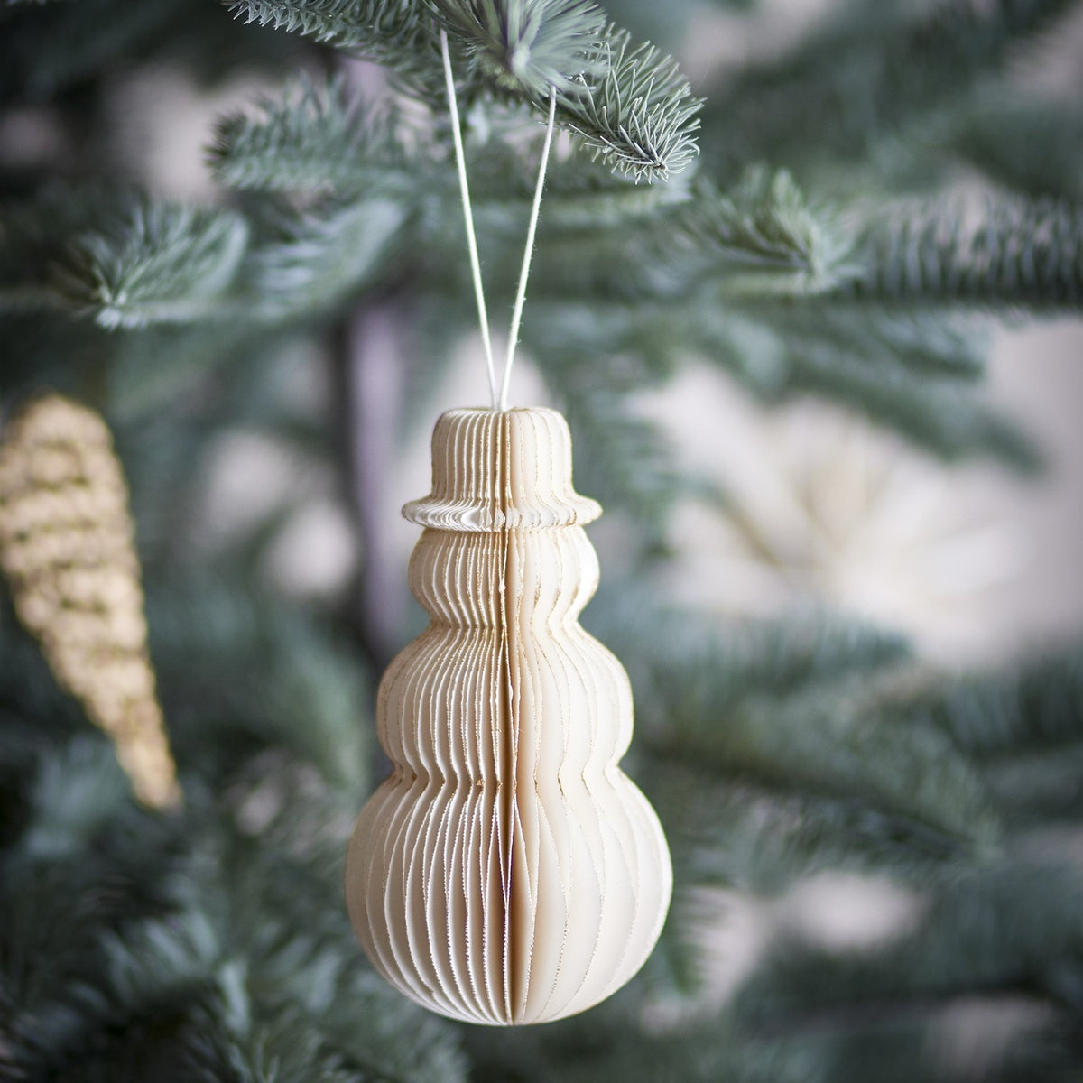 Felip Paper Christmas Tree Decoration in cream, a honeycomb snowman ornament with brimmed hat, hanging by string from a pine tree branch.