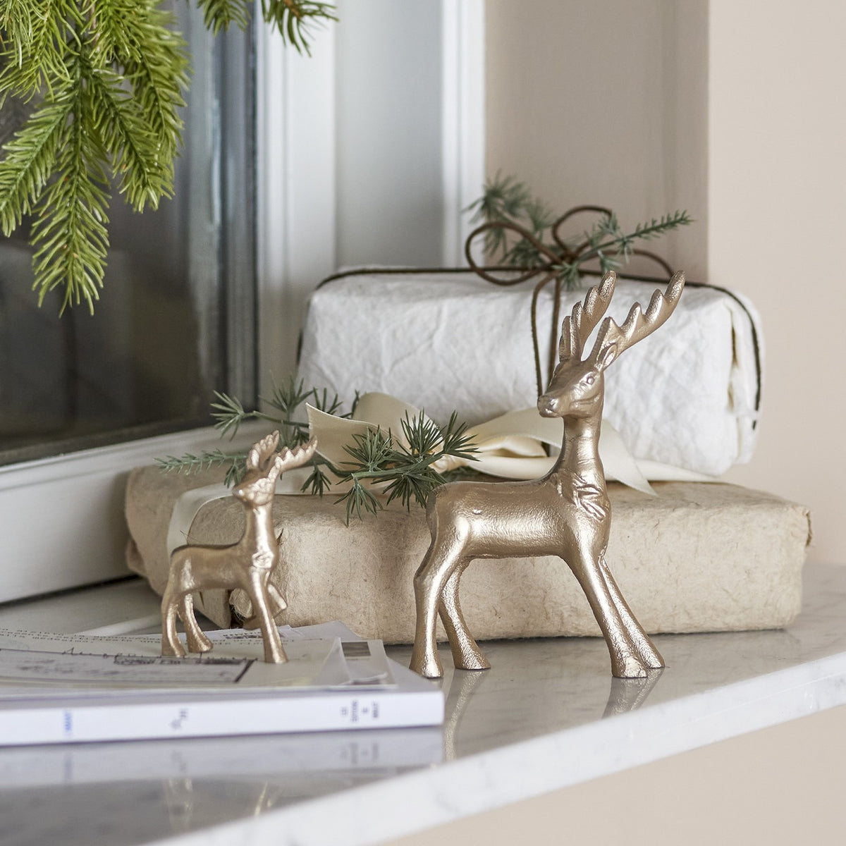 Kila Aluminium Christmas Decorations Set of 2 in gold, featuring a larger antlered stag and a smaller deer with lifted leg, displayed on a marble shelf with wrapped gifts and pine branches.