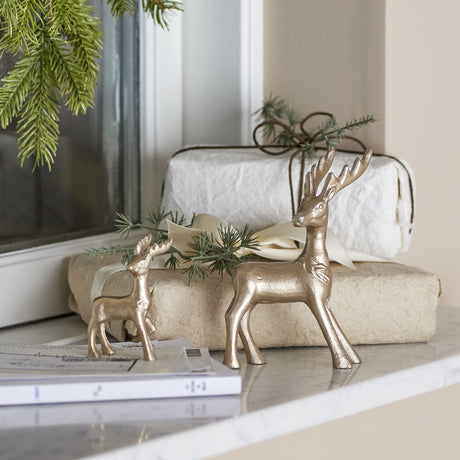 Kila Aluminium Christmas Decorations Set of 2 in gold, featuring a larger antlered stag and a smaller deer with lifted leg, displayed on a marble shelf with wrapped gifts and pine branches.