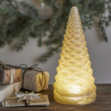 Emilius embossed glass Christmas tree decoration in white, tiered cone shape lit from within, displayed on a wooden surface with gift boxes and festive foliage.