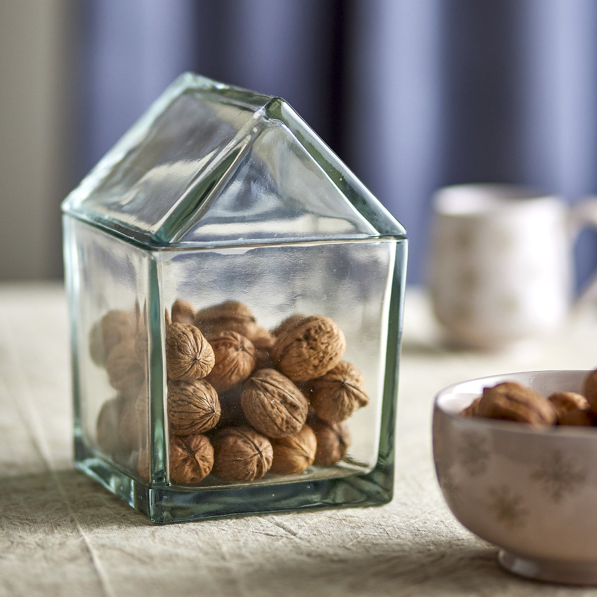 Elius house-shaped clear glass storage jar with pitched roof lid, filled with walnuts, shown on a table beside a bowl of nuts and a mug.