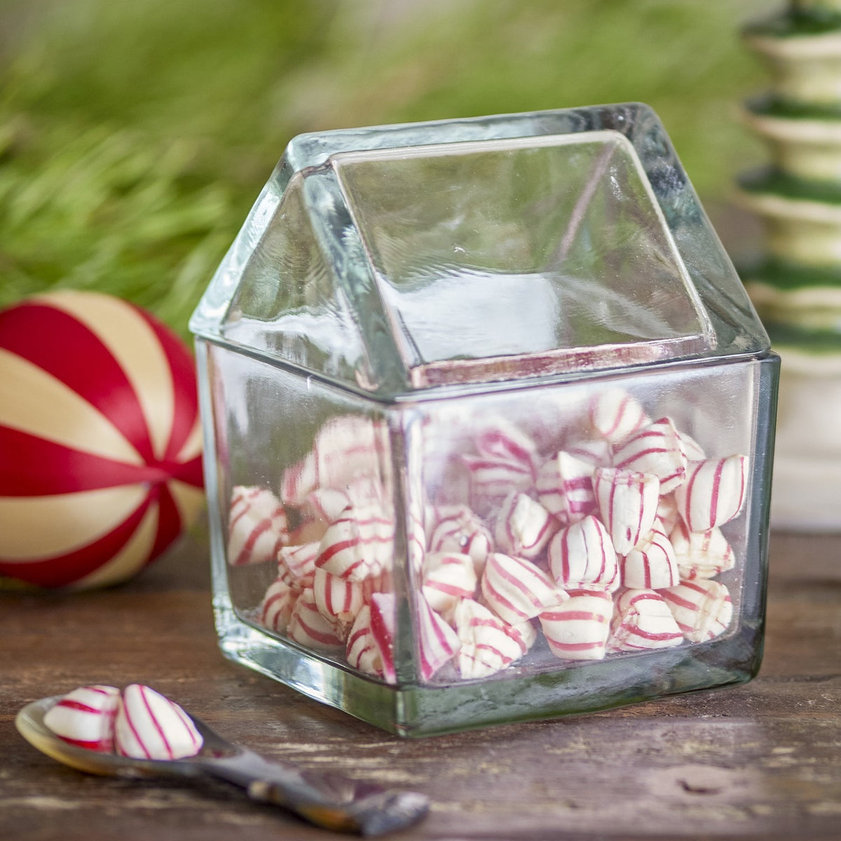 Elius House clear glass storage jar with pitched roof-shaped lid, filled with striped peppermint sweets, shown on a wooden surface with festive decorations.